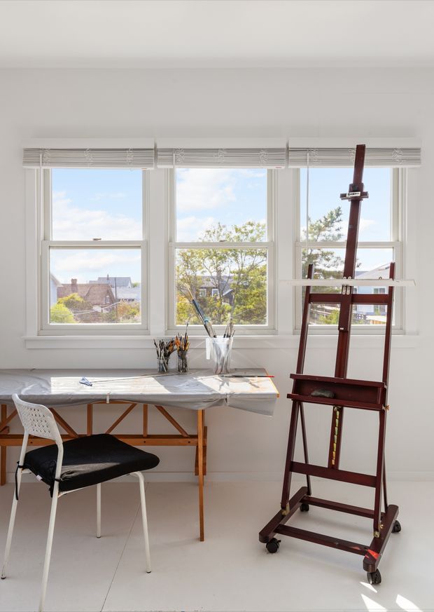Bright, minimalist artist's studio or workspace with three large windows, white walls and flooring, a simple worktable covered in drawing materials, a chair, and a tall dark wood easel. Daylight and residential rooftops are visible outside.