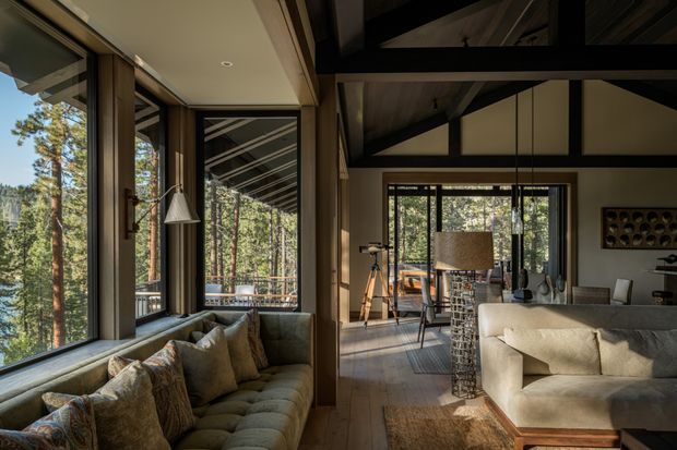 A warm, rustic-contemporary living room in a forest setting, featuring extensive floor-to-ceiling glazing, a built-in upholstered window seat, a large sofa, timber flooring, exposed dark timber roof beams, and a dining area beyond. Tall pine trees are visible through the windows and reflected glazing.