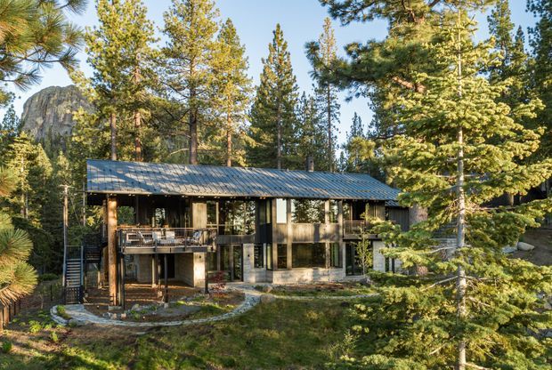 A modern two-storey cabin-style home set among tall pine trees in a forested mountain setting. The building features a long low-pitched metal roof, extensive glazing, dark timber or metal cladding, elevated decks, and a lower-level terrace. A rocky mountain outcrop is visible behind the trees.