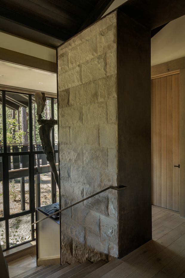 Interior view of a stairway beside a tall, rough-faced natural stone feature wall and dark timber-clad structural volume. A slim metal handrail follows the stairs, with full-height black-framed glazing opening toward a wooded exterior and vertical timber doors to the right. The space has a warm, moody, materially rich contemporary-rustic character.