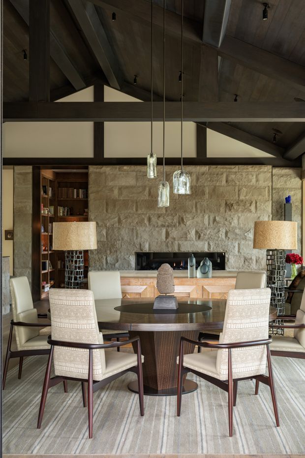 A refined dining room with a large round timber dining table and upholstered mid-century-inspired chairs, set before a rustic stacked-stone fireplace wall. The room features exposed dark timber beams and a timber-lined vaulted ceiling, pendant glass lights, built-in shelving, table lamps, and a neutral striped rug. The overall atmosphere is warm, sophisticated, and lodge-like.