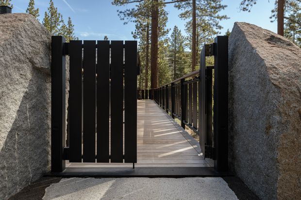 A contemporary outdoor walkway or bridge passing between large natural granite boulders. A black vertical-slat metal gate and matching railing frame the timber-toned deck, with pine trees visible beyond.