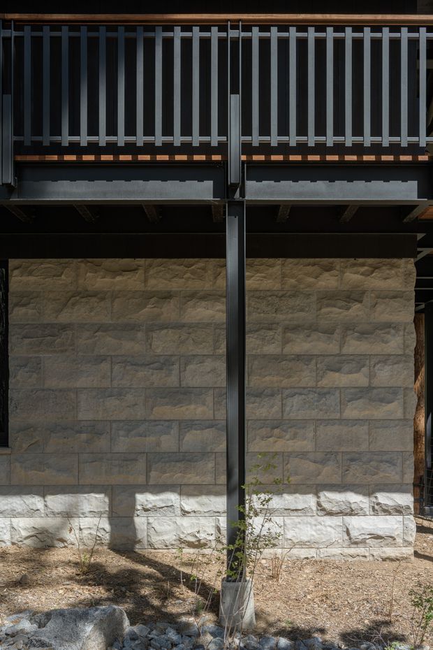 Close-up exterior architectural view of a dark metal-framed facade with a strong vertical structural post, upper screening or balustrade formed by closely spaced vertical metal slats, exposed timber elements, and a lower wall clad in large rough-cut natural stone blocks. The base includes gravel, rocks, and sparse landscaping. The composition is industrial and textural, with strong contrast between the dark structure and warm grey stone.
