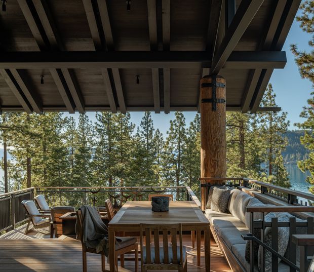 Covered outdoor dining and lounge terrace on a timber deck, with a large solid timber support post, exposed dark roof structure, built-in cushioned bench seating, dining table and chairs. The terrace overlooks a wooded mountain landscape and lake or valley beyond.