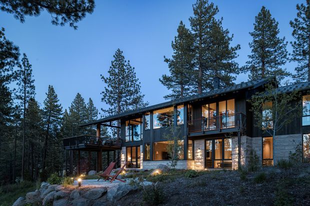 A contemporary multi-level home nestled among tall pine trees on a rocky, wooded site. The dark-clad facade features extensive floor-to-ceiling glazing, projecting balconies, warm interior lighting, and a low-sloped roofline, creating a strong connection to the forest landscape.