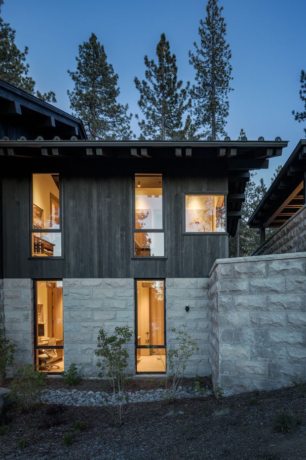 Exterior view of a contemporary multi-level house set among tall pine trees. The facade combines dark vertical timber cladding with large black-framed windows and a lower level faced in rough-cut grey stone. Deep roof overhangs with exposed eaves and layered roof forms create strong horizontal lines, while warm interior lighting glows through the glazing. A stone retaining wall and landscaped gravel garden sit in the foreground.