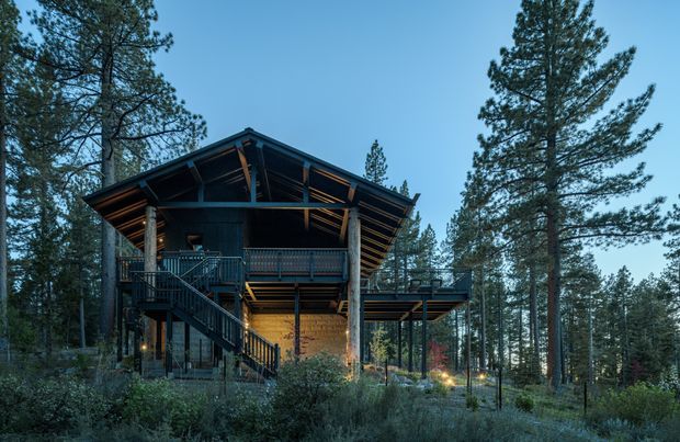 Exterior view of a multi-level timber cabin or lodge set among dense pine forest. The building has a prominent pitched gable roof with exposed structural framing, dark wood cladding, elevated decks and balconies, exterior stairs, and warm interior and landscape lighting visible at dusk.