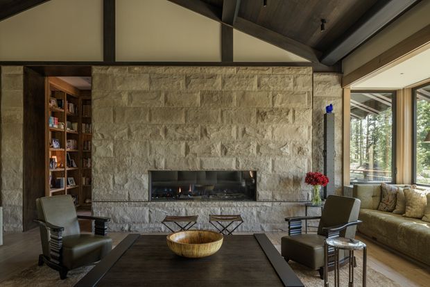 A refined living room centred on a large rough-cut stone fireplace wall with a linear firebox. The room has a high vaulted timber-lined ceiling with exposed dark structural beams, built-in timber shelving, dark wood flooring, leather lounge chairs, a low coffee table, and a large upholstered window seat or sofa beside expansive glazing overlooking trees.