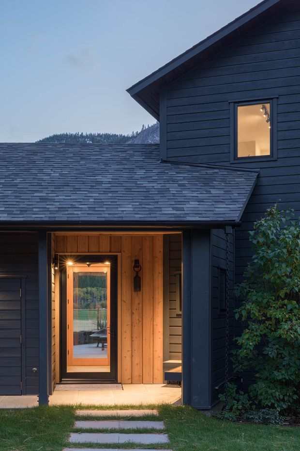 Twilight exterior view of a dark-clad residential house entrance. The facade features horizontal dark timber weatherboards, a steep gabled roof with grey shingle roofing, and a warmly lit recessed entry clad in natural timber. A glazed front door opens toward a small interior stairway, while a stepping-stone path crosses the lawn to the entrance. A forested landscape is visible in the background.