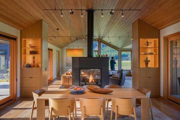 Warm, open-plan dining and living space with a large timber dining table and chairs in the foreground, flanked by built-in timber cabinetry and display shelving. A central double-sided fireplace separates the dining area from a lounge with generous glazing and views to the landscape. The room features extensive timber lining to the ceiling and walls, exposed dark structural elements, contemporary track lighting, and a strong connection to the outdoors.