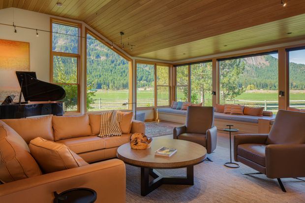 Warm, expansive living room with full-height corner glazing overlooking a rural mountain landscape. The room features a timber-lined ceiling, extensive timber-framed windows, tan leather sofas and armchairs, a round coffee table, a grand piano, and built-in window seating. The palette is earthy and inviting, with abundant natural light and strong indoor–outdoor connection.