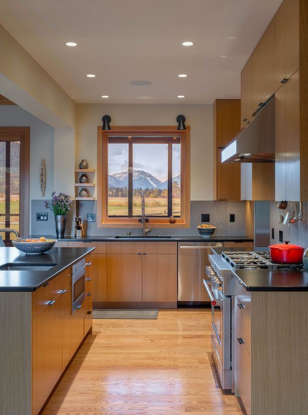 A warm, well-appointed galley-style kitchen with extensive mid-tone timber cabinetry, dark stone benchtops, stainless-steel appliances, and a central island. A large timber-framed window above the sink overlooks a mountain landscape, while recessed ceiling lights provide even illumination. Open shelving, a dining or living area glimpse, and kitchen utensils add lived-in detail.