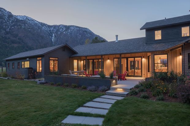 Exterior dusk view of a contemporary mountain home with dark horizontal cladding, multiple gabled roof volumes, warm illuminated glazing, and a covered outdoor terrace. A landscaped lawn and stepping-stone path lead toward the entrance, with a dramatic forested mountain backdrop.