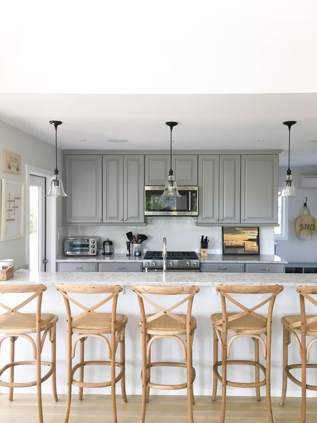 Bright, spacious kitchen with a long white island and five light timber rattan cross-back bar stools. The kitchen features extensive grey Shaker-style cabinetry, white benchtops and backsplash, integrated appliances, a freestanding stainless-steel range, pendant lights, and pale timber flooring. The overall look is clean, warm, and lightly coastal.