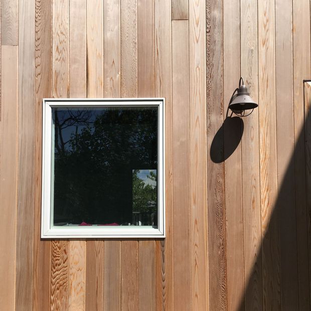 Close-up exterior view of a vertically clad timber wall with varied natural grain, a white-framed window, and a black gooseneck wall light. Strong sunlight creates shadows across the warm wood facade; the window reflects surrounding trees.