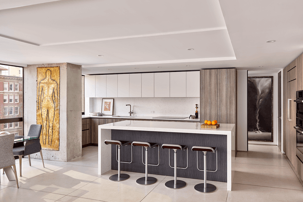 A bright, open-plan contemporary kitchen and dining area. The kitchen features a large island with four bar stools, white upper cabinetry, dark textured lower cabinetry, a pale stone benchtop, integrated appliances, and a minimalist linear splashback. A concrete-look structural column with artwork, a dining table, large windows, and a hallway with timber-toned wall panelling are also visible.