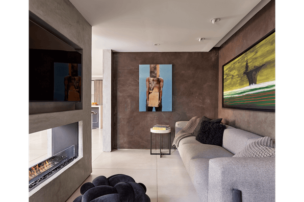 Contemporary living room with a large light-grey sectional sofa, dark textured feature walls, polished pale flooring, recessed ceiling lights, abstract artwork, and a sculptural dark ottoman. A double-sided or built-in fireplace and wall-mounted television occupy the left side, while a glimpse of the adjacent kitchen or hallway is visible beyond the room.