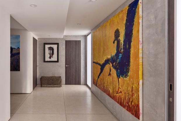 A contemporary residential hallway with large-format pale tiled flooring, smooth light walls, recessed ceiling lighting, and dark wood-grain internal doors. A dramatic oversized abstract artwork dominates the right wall, while a small woven bench and framed artwork sit near the end of the corridor.