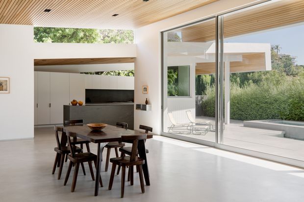 Bright contemporary open-plan dining and living space with a timber slatted ceiling, smooth pale flooring, white built-in cabinetry, and full-height glazed sliding doors opening to a landscaped outdoor terrace. A dark timber dining table and chairs occupy the foreground, while a compact living area and television are set into the rear joinery. The glazing provides views into a modern courtyard-like exterior with matching timber soffits and planted screening.