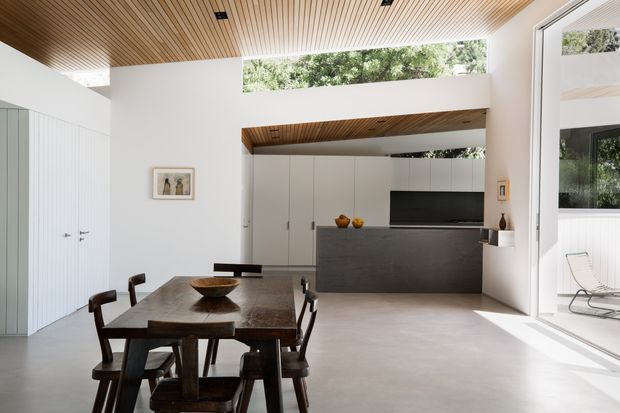 A bright, open-plan contemporary dining and living area with a large dark timber dining table and chairs in the foreground. White built-in cabinetry forms the kitchen or media wall, with a dark grey island or counter, integrated television, and minimal decor. The room features a warm timber slatted ceiling, high clerestory glazing, large sliding glass doors, and a light polished floor, creating a strong connection to the outdoor patio.