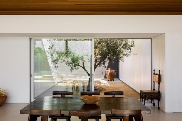 A refined dining room with a substantial dark timber dining table and matching benches and chairs, centred beneath a warm timber-lined ceiling. Full-height glazing opens the room visually toward a landscaped courtyard, while white walls, vertical wall panelling, and a large earthenware bowl create a calm, natural interior. A narrow timber console and decorative objects sit along the right wall.