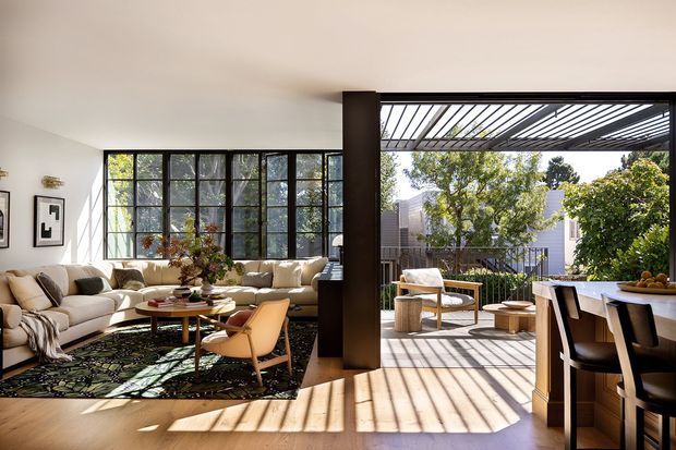 A bright, open-plan contemporary living and dining area connected to a covered outdoor terrace. The living room features large black-framed steel-look windows, a cream sectional sofa, patterned rug, timber coffee tables, and a dark structural column. The terrace has a slatted pergola, outdoor lounge seating, and a dining counter with stools, with abundant greenery beyond.