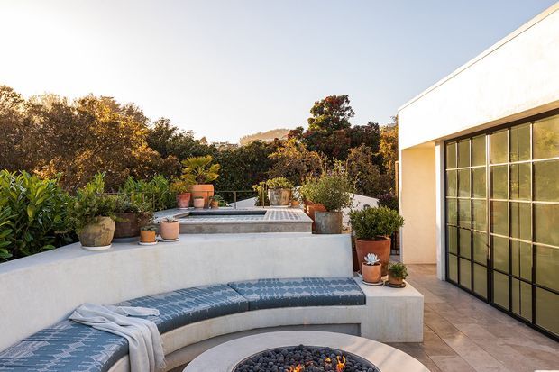 A sunlit contemporary outdoor terrace with a curved built-in white rendered bench surrounding a round fire pit, patterned cushions, and potted plants. A raised rectangular water feature or plunge pool sits behind the seating, while a large black-framed glazed door opens onto the terrace. The setting includes mature landscaping and a low hillside in the background.