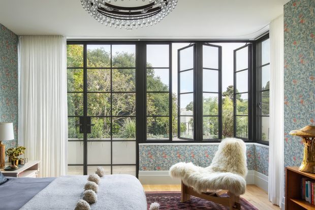 Bright, refined bedroom with a bed in the foreground, botanical patterned wallpaper, pale timber flooring, white curtains, and a large black-framed bay of divided-light windows opening outward to a leafy view. A sculptural chandelier, fur-covered lounge chair, bedside table, and timber shelving add layered decorative detail. The room has an eclectic, elegant character with strong natural light and garden outlook.