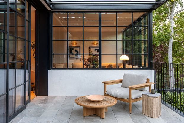 A modern glazed outdoor terrace or sunroom adjoining a warmly lit interior. Black metal-framed floor-to-ceiling glazing and a glass roof create a strong indoor-outdoor connection, while pale rendered walls, large grey floor tiles, timber lounge furniture, and black metal balustrading give the space a refined contemporary character. Trees are visible beyond the railing.