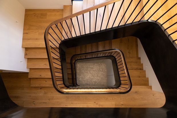 Top-down view through a multi-level stairwell with a geometrically curved stair and landing arrangement. Warm timber stair treads and landings contrast with dark metal balustrades and a dark floor edge. The repeated rectangular opening creates a strong sculptural spiral effect, with white plaster walls and a patterned surface visible at the bottom of the stairwell.