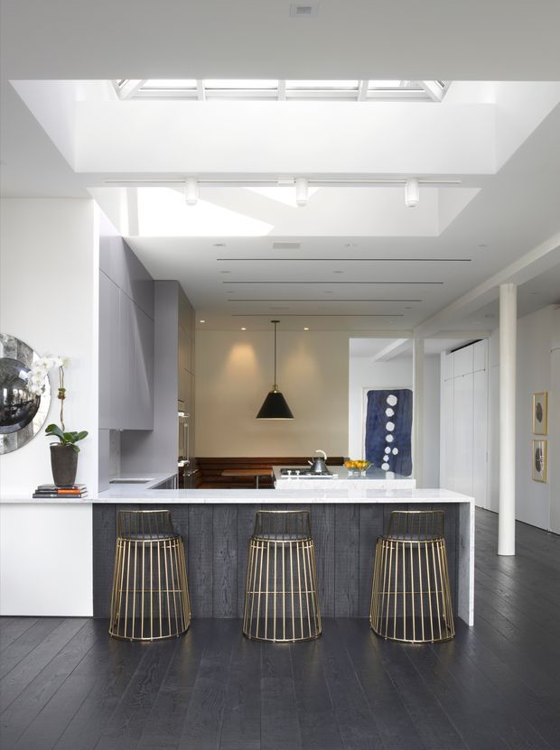 Bright contemporary kitchen interior with a large central island, dark textured cabinetry, white stone benchtop, and three brass-toned bar stools. The space features dark timber flooring, white walls and joinery, recessed linear lighting, skylights, and a sculptural black pendant over the rear dining area. A hallway and artwork are visible beyond the kitchen.