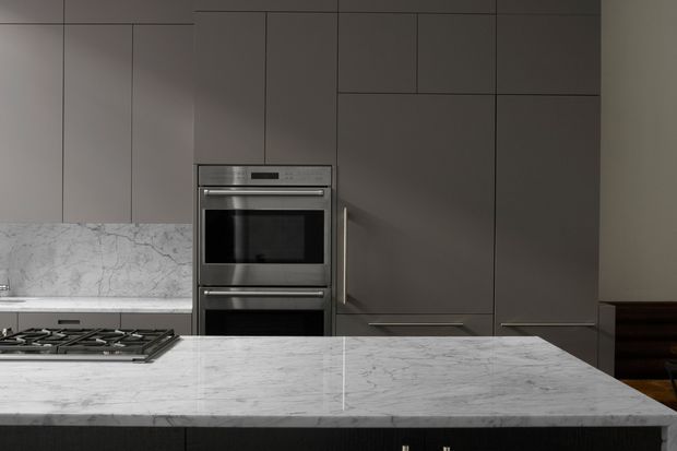 A refined contemporary kitchen viewed across a large marble island. Floor-to-ceiling dark taupe cabinetry forms a minimalist wall with integrated stainless-steel double ovens, while a pale veined marble splashback and island benchtop provide contrast. A gas cooktop is set into the island, and the room has a restrained, tonal, high-end finish.