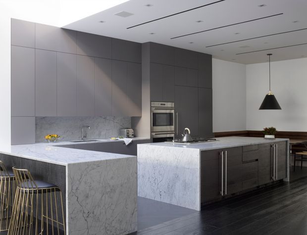 A spacious contemporary kitchen with a large marble-clad island and waterfall benchtop, a separate peninsula with gold-framed bar stools, matte dark grey full-height cabinetry, integrated stainless-steel appliances, and dark timber flooring. A dining area is visible beyond the kitchen, with minimalist recessed ceiling lighting and a black pendant light.