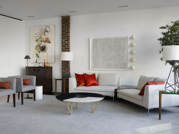 A bright, refined living room with a neutral grey and white palette, arranged with a sofa, armchairs, a chaise-style lounge, and layered round coffee tables. Red cushions provide bold accents. The room features a white ceiling with recessed lighting, pale carpet, a dark timber sideboard, a narrow exposed brick column, contemporary artwork, sculptural wall decor, and indoor greenery.