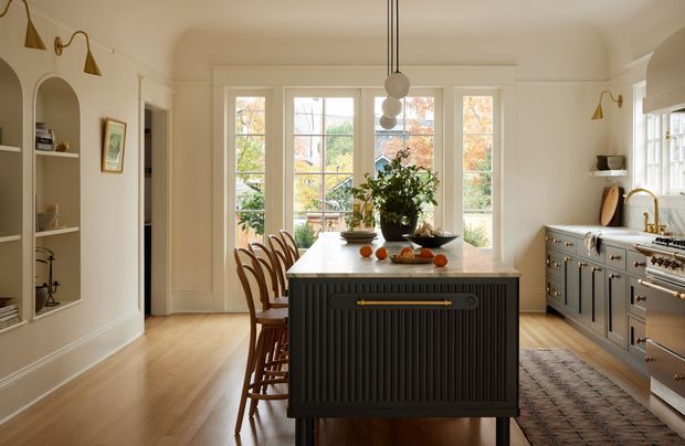 A bright, refined kitchen with a large central island featuring dark fluted cabinetry and brass hardware, surrounded by warm timber flooring, pale cabinetry, integrated shelving, and a freestanding range. Tall multi-pane windows and glazed doors bring in abundant natural light, while pendant globes hang above the island. A patterned runner, timber bar stools, and decorative objects add warmth and character.