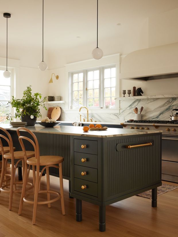 A warm, refined kitchen centred on a large dark fluted island with brass hardware and a pale stone benchtop. Wooden counter stools sit along the island, while a large multi-pane window, veined marble splashback, range cooker, open shelving, pendant lights and timber flooring define the room. The palette combines cream and white surfaces, dark cabinetry, natural timber and brass accents.