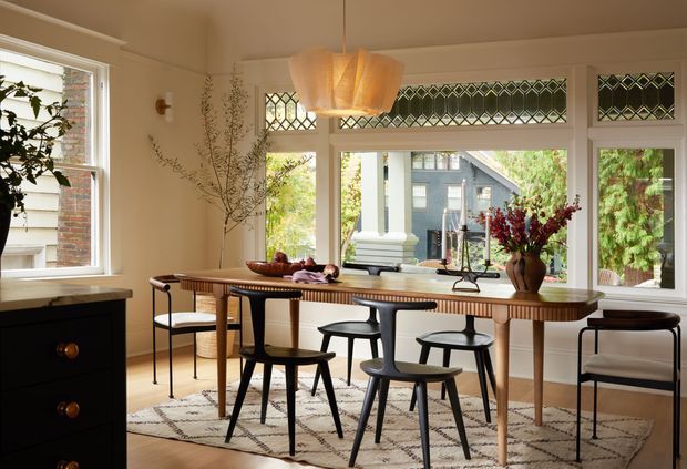 A warm, refined dining room with a long timber dining table and mid-century-style black chairs arranged on a patterned rug. Large windows and decorative diamond-patterned transom glazing provide abundant natural light and views of greenery and neighbouring homes. A sculptural pendant light, floral arrangement, and dark built-in cabinetry complete the inviting interior.