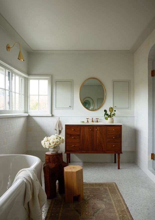 A bright, refined bathroom featuring a freestanding white bathtub, a timber mid-century-style vanity with brass fixtures, white tiled walls, patterned floor tile, an oval mirror, and large divided-light windows. Warm wood furniture, brass accents, and floral styling create an elegant traditional-contemporary atmosphere.