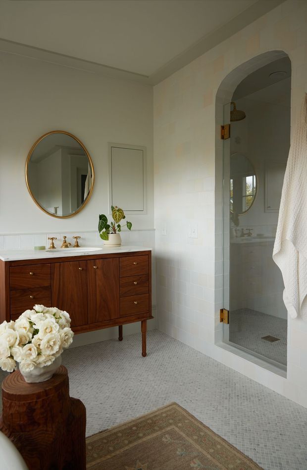 A refined bathroom with a floating-style dark timber vanity, white stone benchtop, paired brass taps, round brass-framed mirror, and a glass-enclosed walk-in shower. The room features pale tiled walls and small-format light mosaic floor tiles, with an arched shower opening, white towels, a potted plant, and warm natural styling.