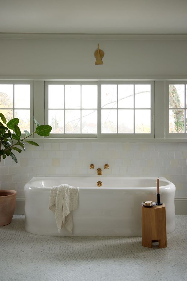 Bright, serene bathroom featuring a freestanding white bathtub beneath a wide bank of white-framed multi-pane windows. The walls are finished with white rectangular tile, and the floor has small pale mosaic tiles. Brass/gold tapware and wall sconce details add warmth, while a leafy potted plant, white towel, and small timber stool with candle provide natural accents.