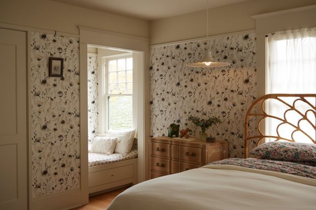 Warm, traditional bedroom with patterned floral wallpaper, a curved rattan or bamboo bedhead, upholstered floral bedding, and a timber chest of drawers. A built-in window seat is visible through the open doorway, while a pendant light and soft daylight create a cosy, vintage atmosphere.