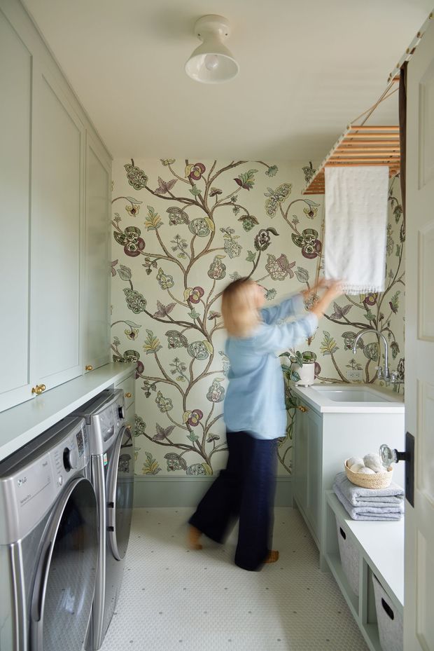 A compact, bright laundry room with a front-loading washer and dryer beneath pale built-in cabinetry on the left, and a white utility sink with chrome tapware on the right. The back wall features ornate botanical and floral wallpaper, while a timber drying rack and folded towels add practical storage. Pale green-grey cabinetry, white surfaces, and patterned flooring create a traditional yet playful interior. A blurred person is reaching toward the drying rack.