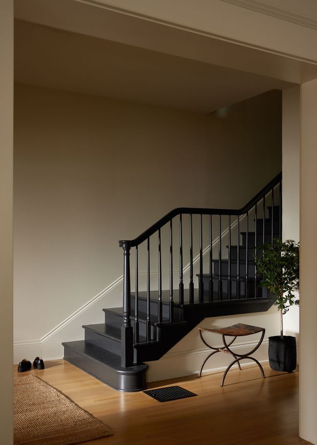 A warmly lit interior stairway with dark painted timber stairs and black turned-newel balustrade, set against beige walls with white trim. Honey-toned timber flooring, a woven rug, a small decorative table, and a potted indoor tree complete the refined traditional setting.