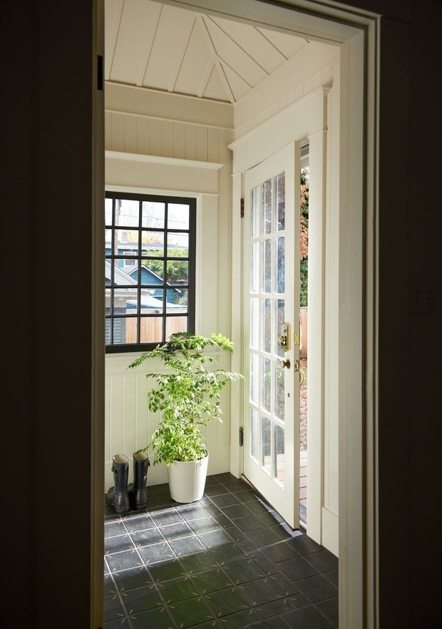 A bright, traditional entry vestibule viewed from an interior doorway. The space features cream-painted vertical wall panelling, ornate trim, a decorative geometric vaulted ceiling treatment, dark patterned tiled flooring, and a glazed white French-style exterior door opening outdoors. A black-framed multi-pane window overlooks neighbouring houses, while a potted plant and boots sit near the entrance.