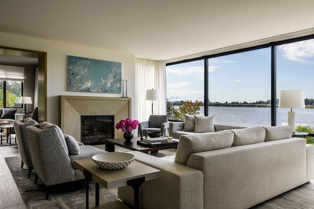 A refined living room overlooking a body of water through expansive floor-to-ceiling glazing. The room features a large neutral upholstered sofa, textured armchairs, a dark coffee table, a stone fireplace surround with a black inset firebox, layered rugs, soft curtains, and understated contemporary furnishings. A dining or adjoining seating area is visible through the opening at left.
