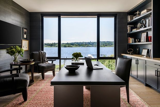 A refined home office with a large dark desk positioned in front of full-height black-framed glazing overlooking a lake. The room features dark textured wall panelling, built-in open shelving and cabinetry, a wall-mounted television or monitor, upholstered chairs, timber flooring, and a patterned rug. The palette is predominantly charcoal, black, grey, and warm timber, creating a sophisticated, moody workspace with strong indoor–outdoor connection.