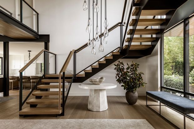 A refined contemporary interior centered on a sculptural open-riser staircase with dark steel framing, warm timber treads, and glass balustrades. A cluster of clear pendant lights hangs over a round stone console table, while full-height glazing, a built-in window seat, indoor planting, and warm timber flooring create a bright, sophisticated entry or living space. A dining area is visible beyond the staircase.