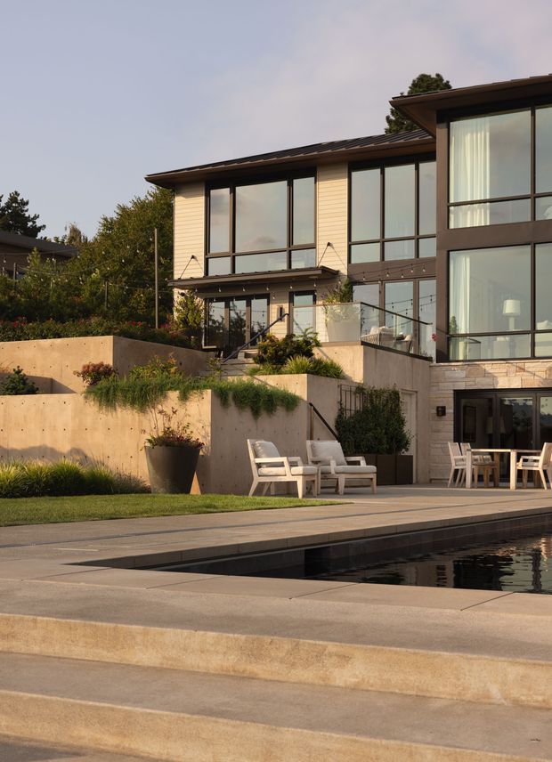 A contemporary multi-level residential house viewed from the pool terrace. The facade combines dark metal-framed floor-to-ceiling glazing, horizontal light cladding, concrete retaining walls, and pale stone masonry. Terraced landscaping, outdoor seating, a paved pool deck, and a long reflecting or swimming pool occupy the foreground.