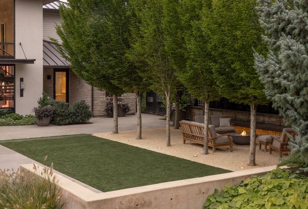 A landscaped residential courtyard viewed from outside, featuring a rectangular lawn enclosed by low concrete retaining walls, rows of mature trees, gravel paving, timber outdoor seating, and a small fire feature. Contemporary rendered and timber-clad building facades are visible behind the garden, creating a private, structured outdoor living space.