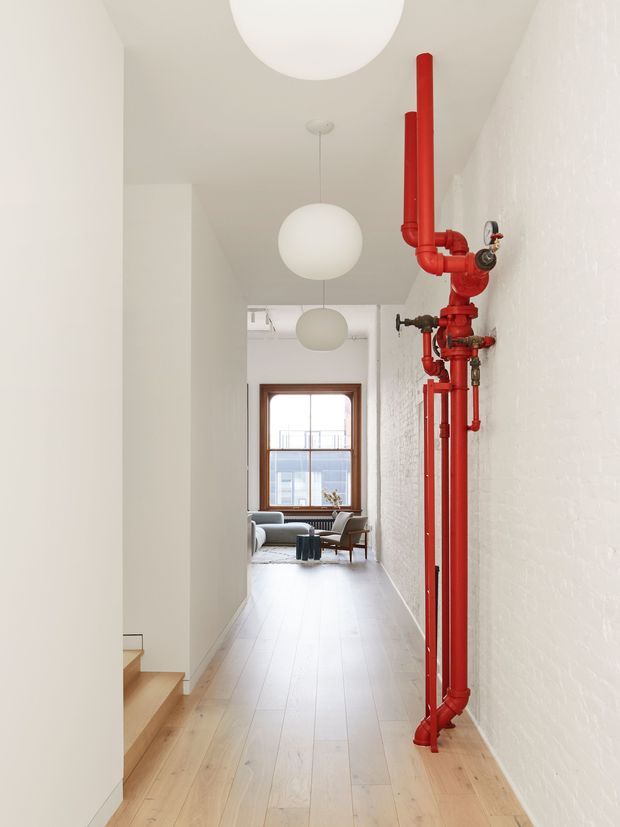Bright, narrow residential hallway with pale timber flooring, white walls and exposed white-painted brick. A vivid red exposed pipe and valve installation runs along the right wall, while spherical pendant lights line the corridor. A short timber stair is visible in the foreground, leading toward a small living area with a window and seating at the far end.